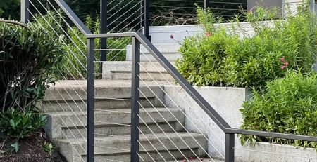 Black metal stair railing with stainless steel cable infill installed on an outdoor concrete staircase with surrounding greenery.