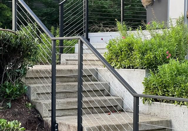 Black metal stair railing with stainless steel cable infill installed on an outdoor concrete staircase with surrounding greenery.