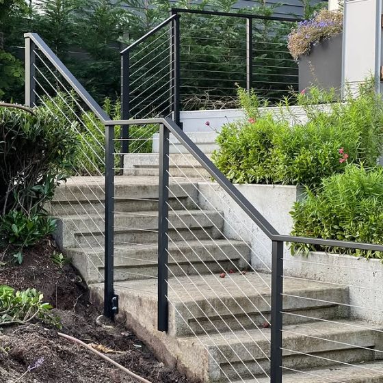 Black metal stair railing with stainless steel cable infill installed on an outdoor concrete staircase with surrounding greenery.
