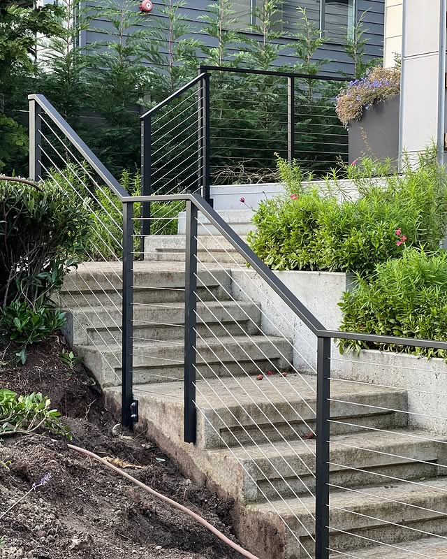 Black metal stair railing with stainless steel cable infill installed on an outdoor concrete staircase with surrounding greenery.