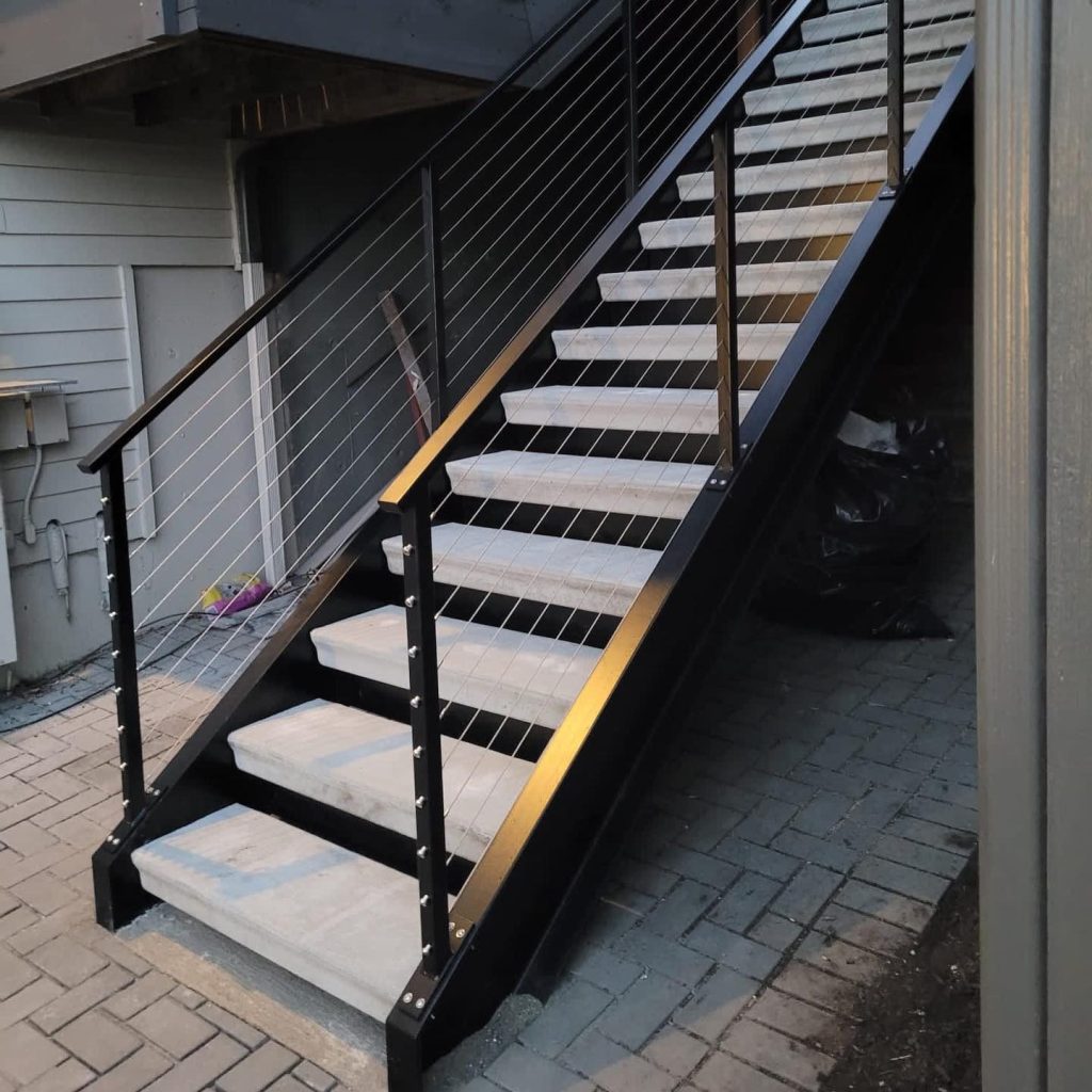 Exterior floating staircase connecting a deck to a patio in Bellevue. Features durable black steel stringers, concrete treads, and a stainless steel cable railing system.